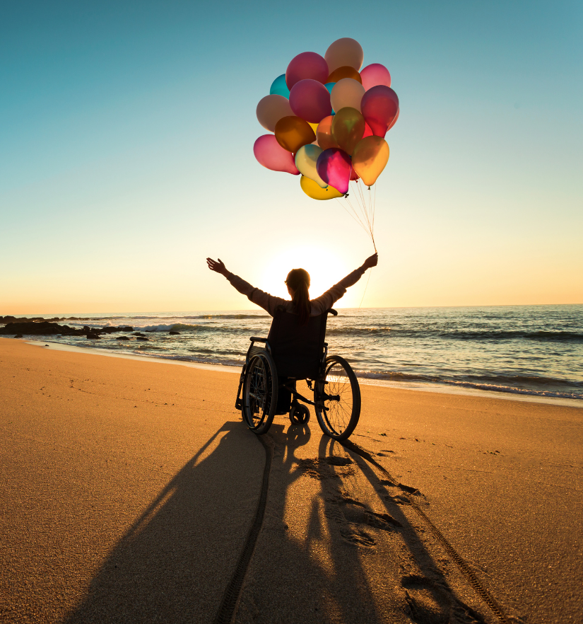 A person in a wheelchair holding colorful balloons on the beach at sunset, celebrating freedom and joy, symbolising YWECARE’s genuine care, commitment, and trusted NDIS support.
