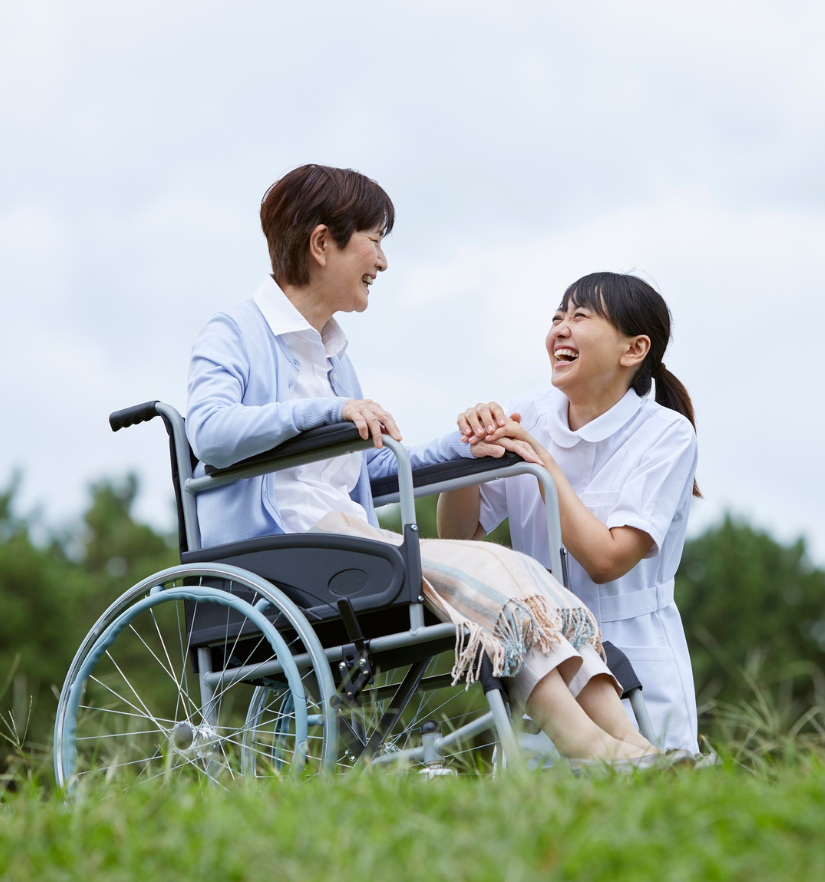 A support worker kneeling beside a smiling woman in a wheelchair outdoors, holding her hands with care and compassion, representing personal care support with YWECARE.