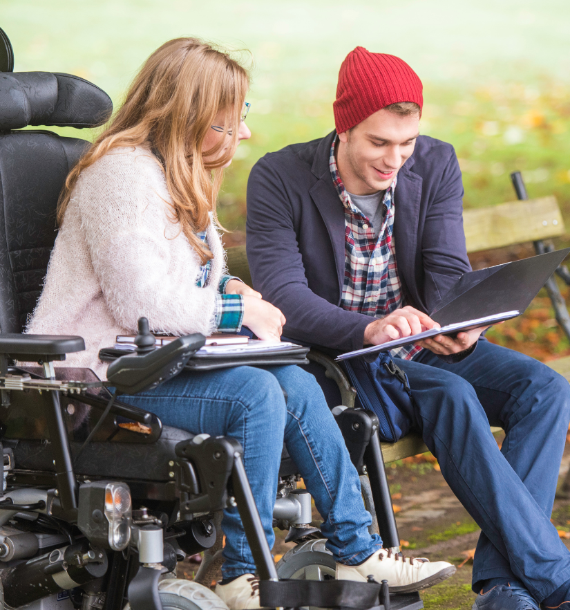 A young woman using a power wheelchair sits with a male support worker on a park bench, both smiling while looking at a laptop, representing NDIS support services and empowerment through YWECARE.