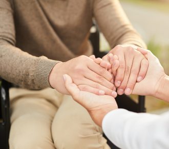 A support worker gently holding the hands of a participant in a wheelchair, symbolising daily living support and compassionate care.
